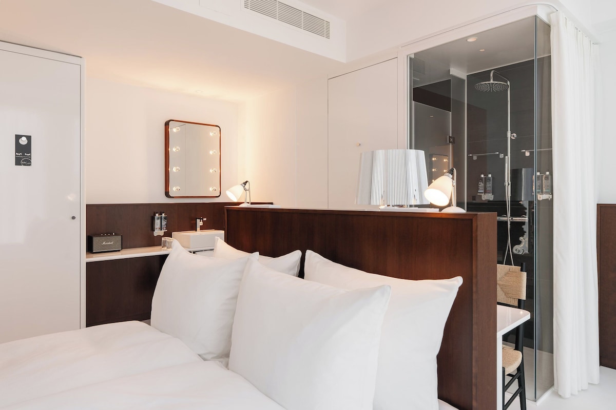 Habitación de hotel con cama, tocador con espejo, zona de ducha y una mesita con lámpara.