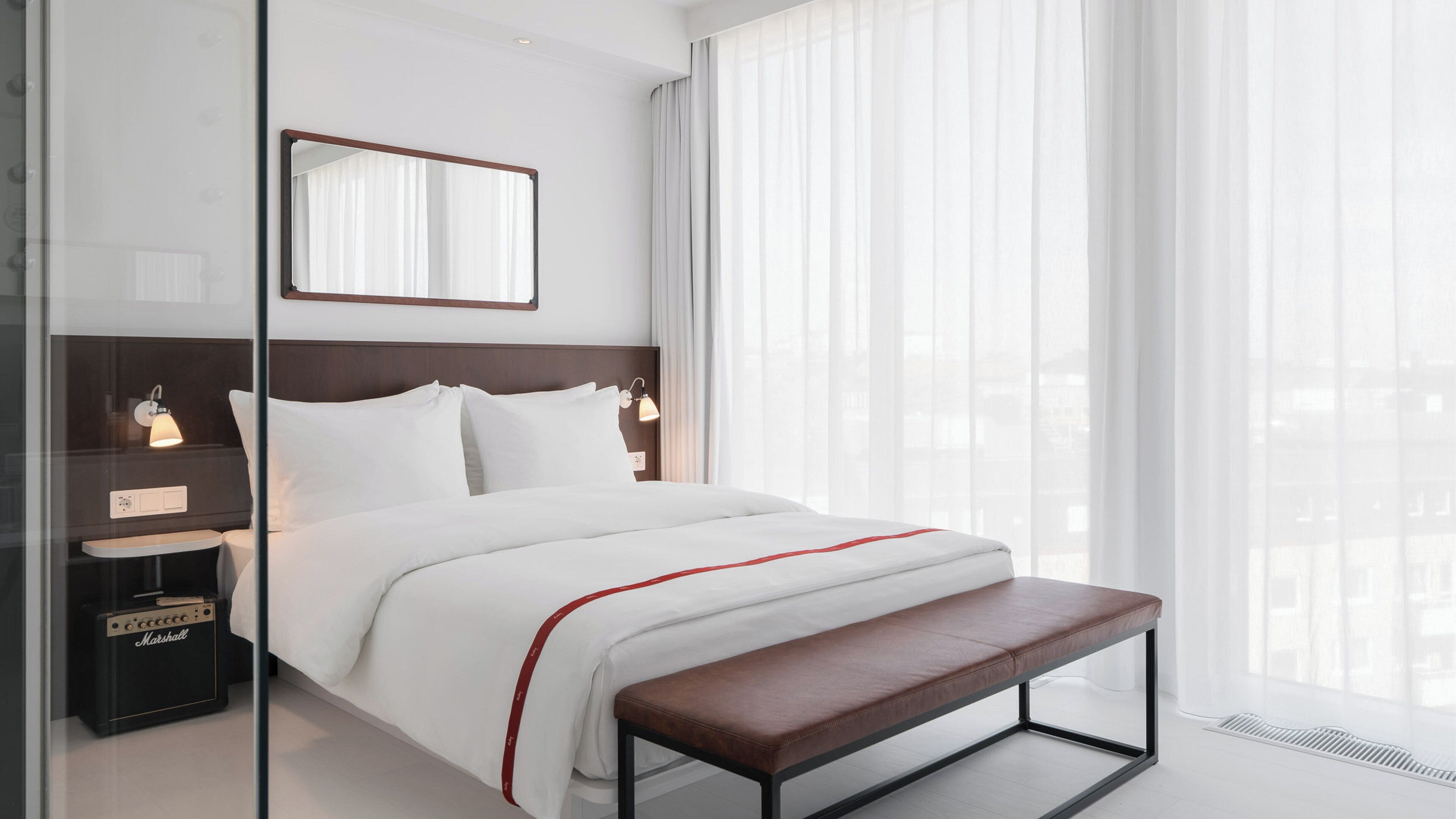 Quarto de hotel moderno com uma cama grande, luminárias de cabeceira, um banquinho e vista através de cortinas finas.