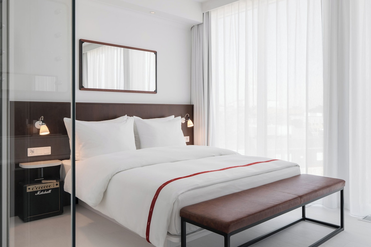 Una habitación de hotel moderna con una cama grande, lámparas de noche, un banco y vistas a través de cortinas traslúcidas.