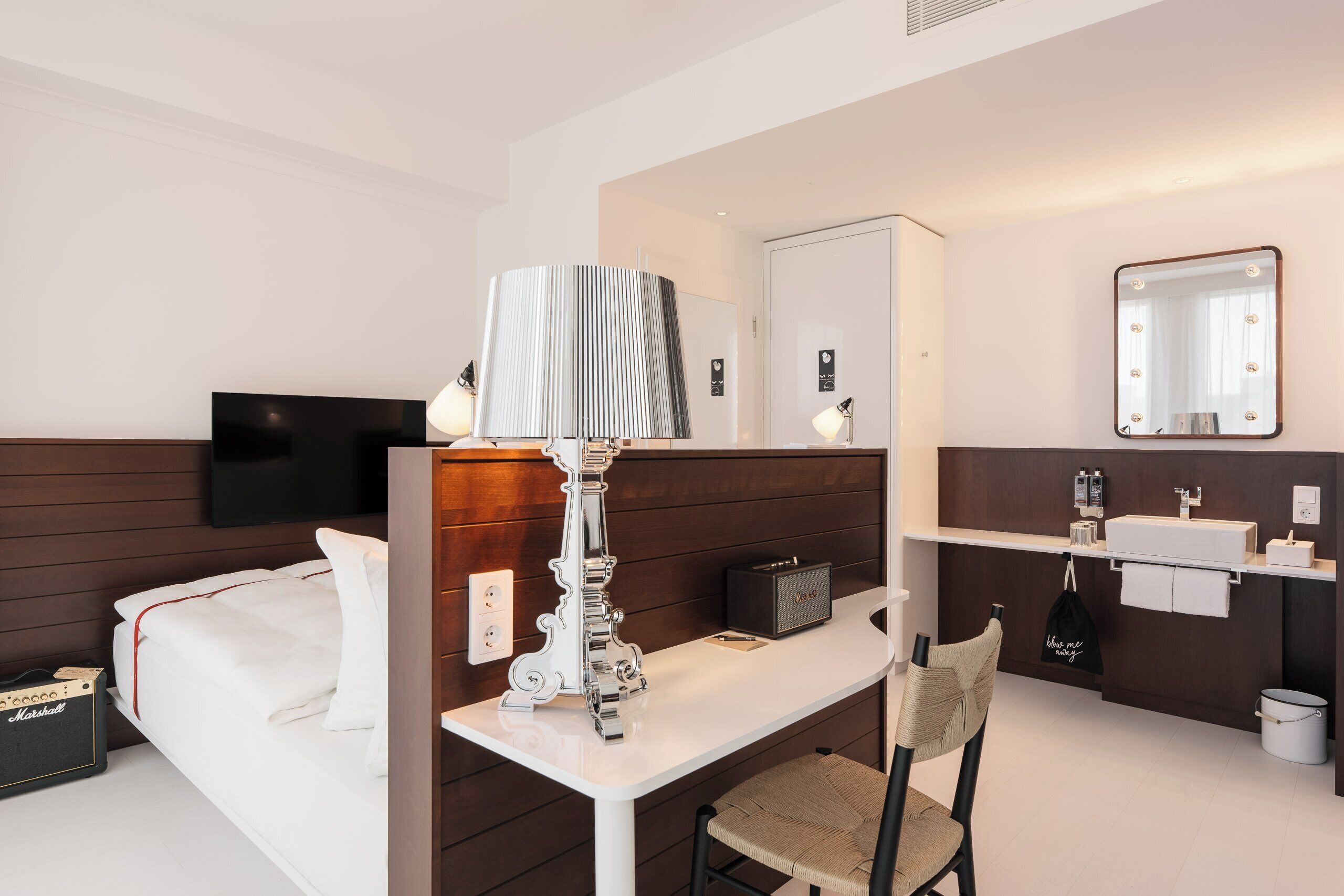 A hotel room with a bed, a desk with a lamp, a chair, a TV, a mirror, a bathroom with a sink, and a trash can.