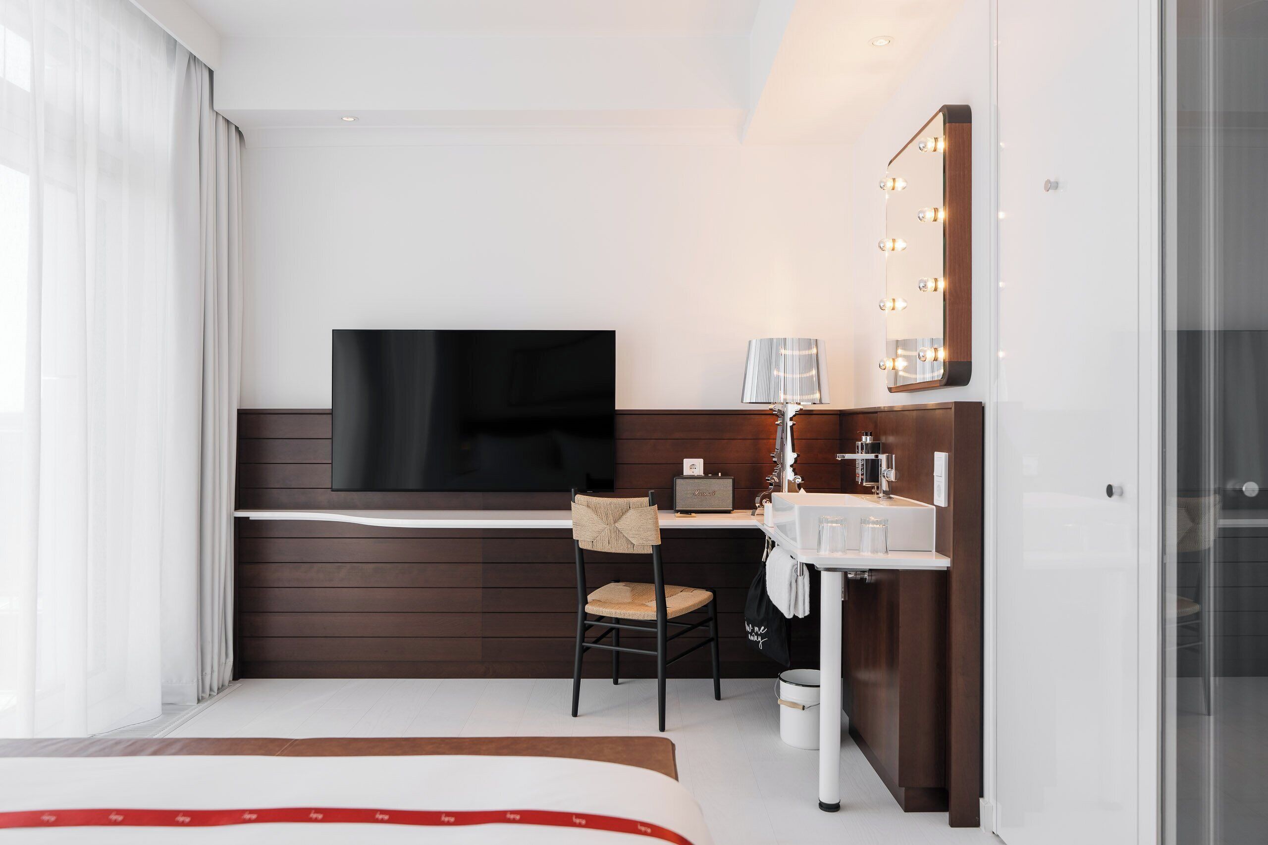 A modern hotel room with a large flat-screen TV, a desk with a chair, a vanity with a mirror and lights, and a bed with a red and white striped bedspread.
