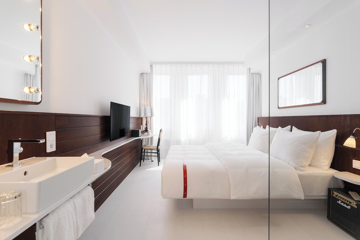 Habitación de hotel con una cama grande, una mesita de noche, un televisor, un espejo, un baño con lavabo y espejo, y una ventana con cortinas traslúcidas.