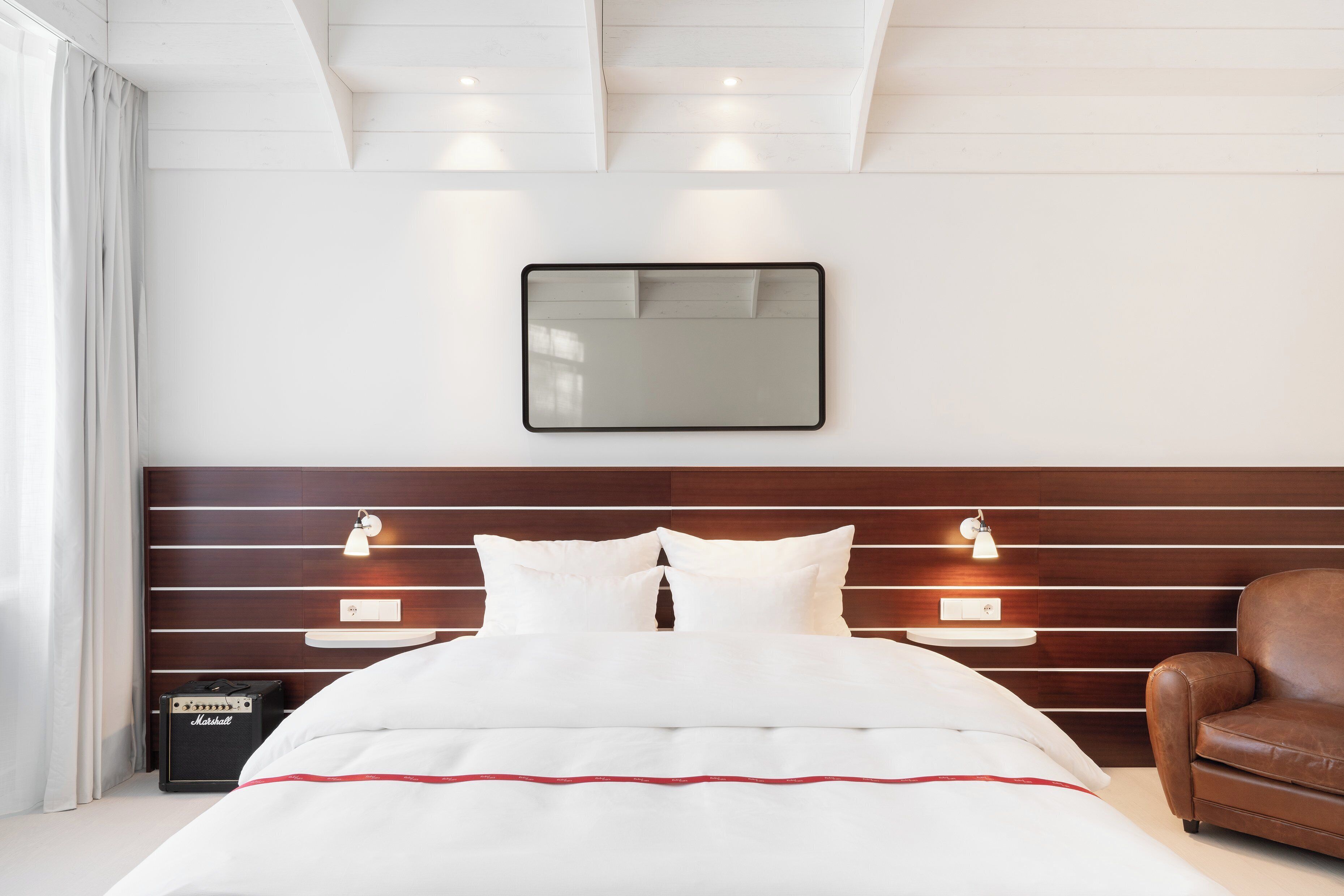 Una habitación de hotel moderna con una cama bien hecha, cabecera de madera, lámparas de noche y un sillón de cuero.
