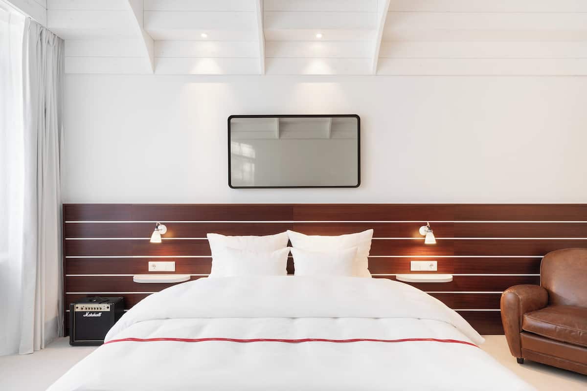 Una habitación de hotel moderna con una cama bien hecha, cabecera de madera, lámparas de noche y un sillón de cuero.