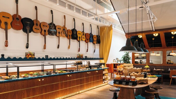 A buffet area with a variety of food items, a wall with hanging guitars, and a seating area with wooden stools. - Ruby Frida Hotel Stockholm By IHG (Stockholm)
