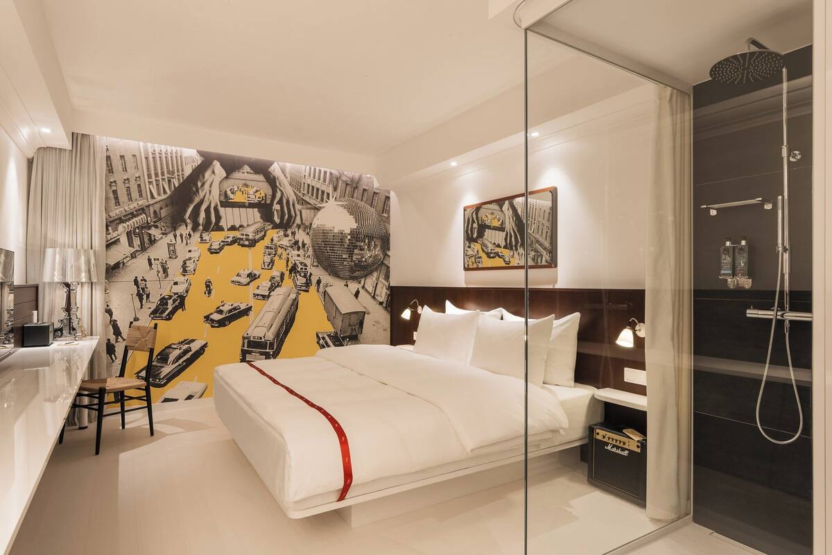 A modern hotel room with a bed, bedside table, lamp, and a glass-enclosed shower area.