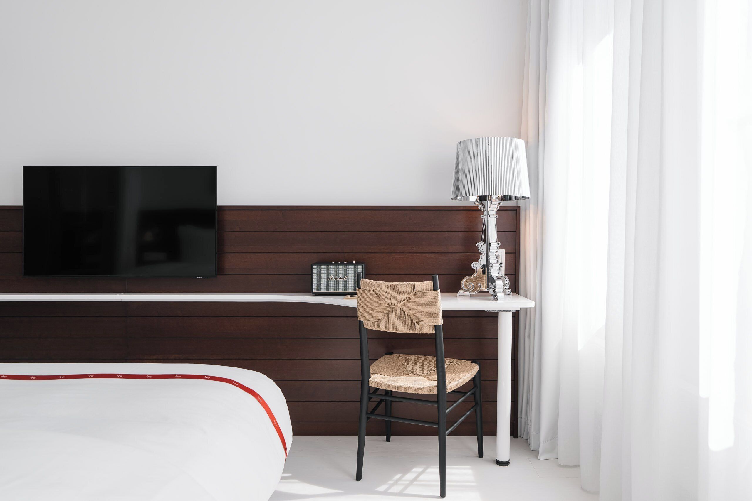 A hotel room with a bed, a desk with a lamp, a chair, and a flat-screen TV.