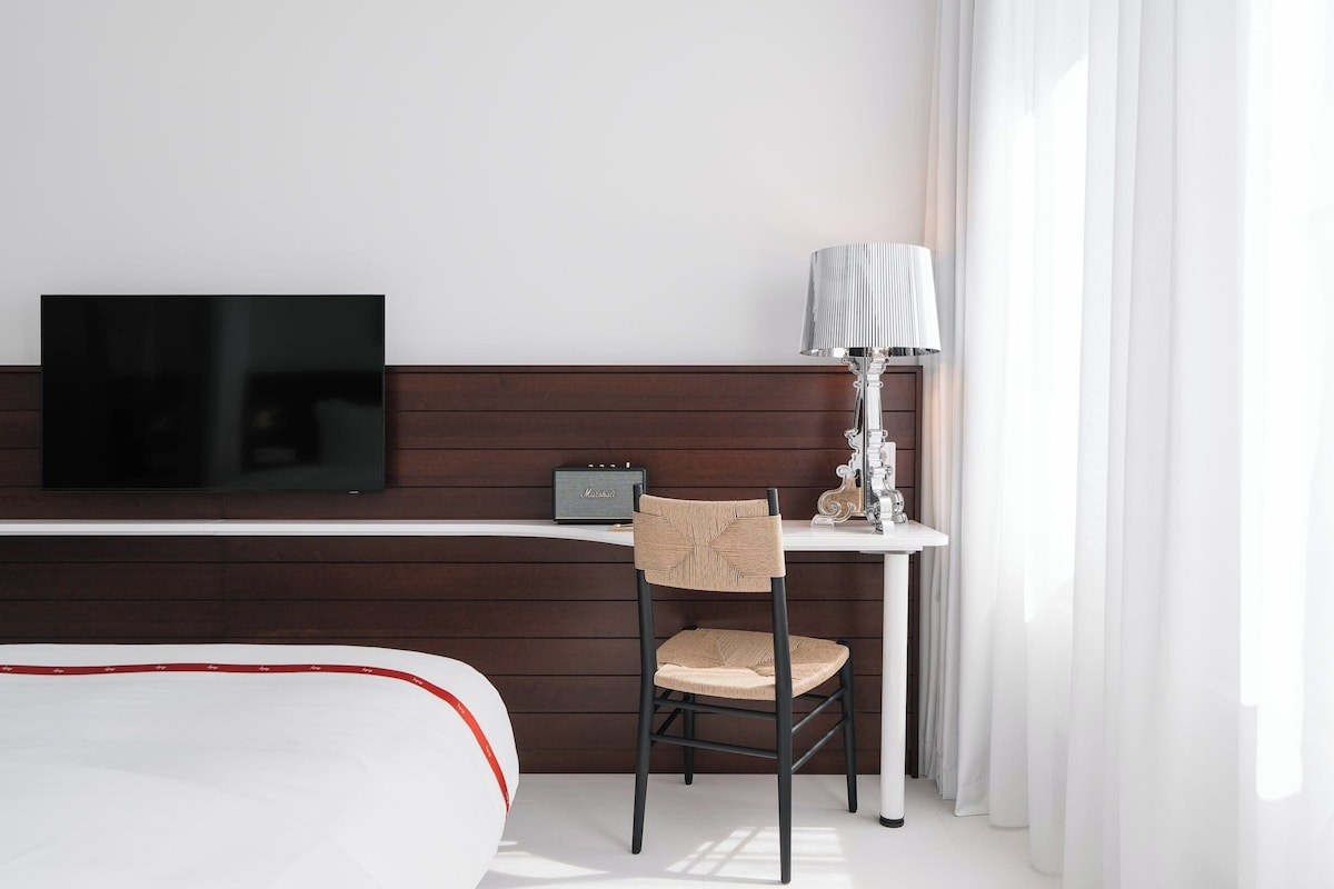 A hotel room with a bed, a desk with a lamp, a chair, and a flat-screen TV.