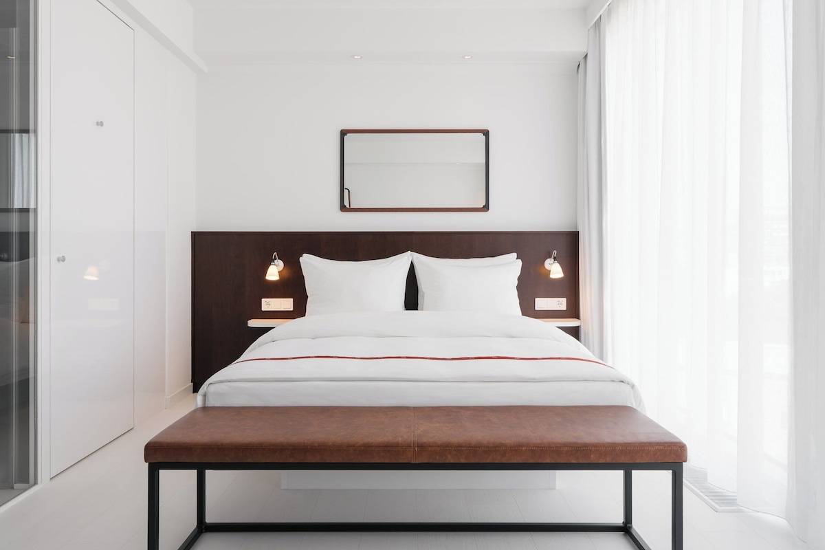 Una habitación de hotel moderna con una cama grande, mesitas de noche, un cabecero de madera, un espejo y una ventana con cortinas.