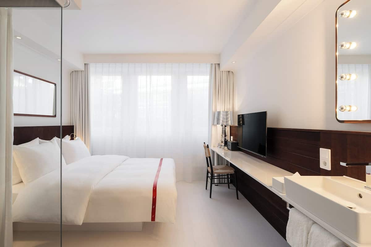 A hotel room with a bed, a desk, a TV, a mirror, and a bathroom.