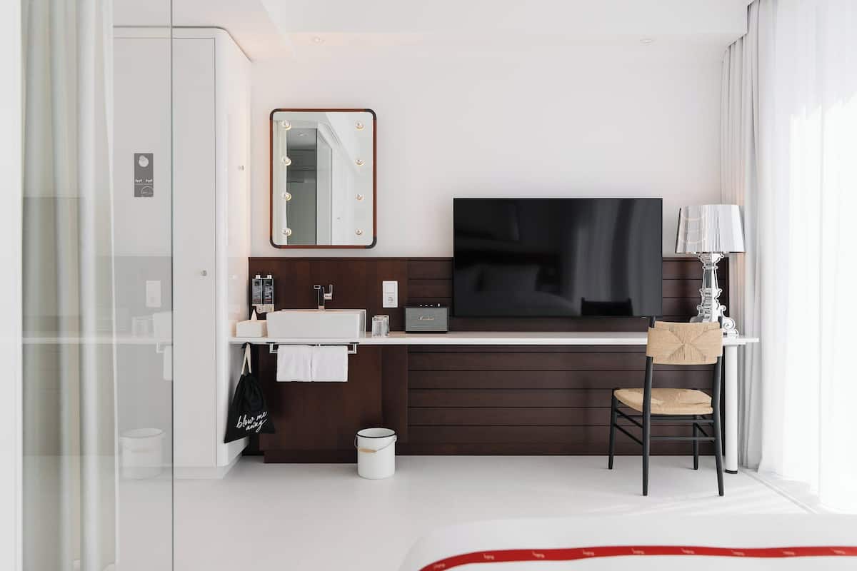 A modern hotel room with a large flat-screen TV, a vanity with a mirror, a chair, and a desk.