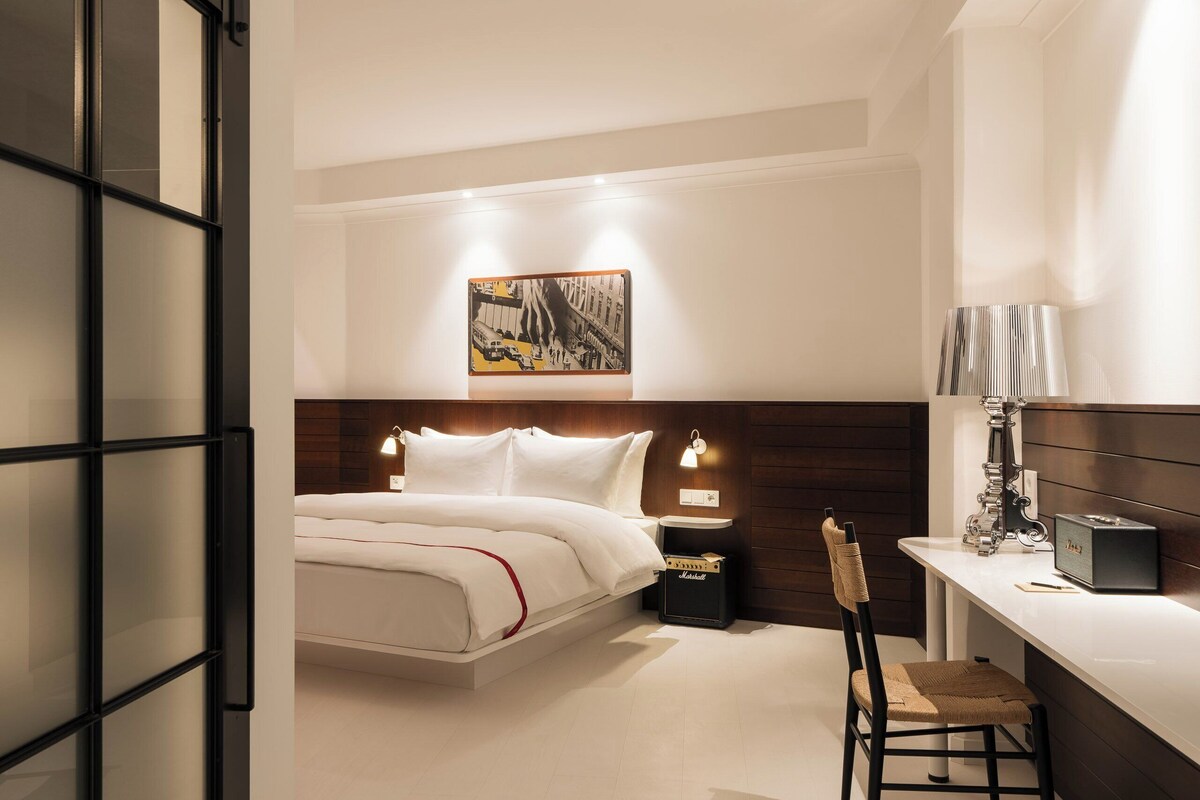 A modern hotel room with a bed, bedside table, lamp, chair, and a painting on the wall.