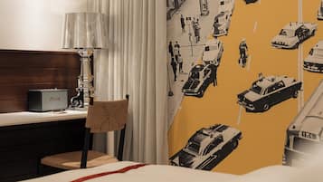 A hotel room with a bed, a desk with a lamp, a chair, and a wall with a car pattern.