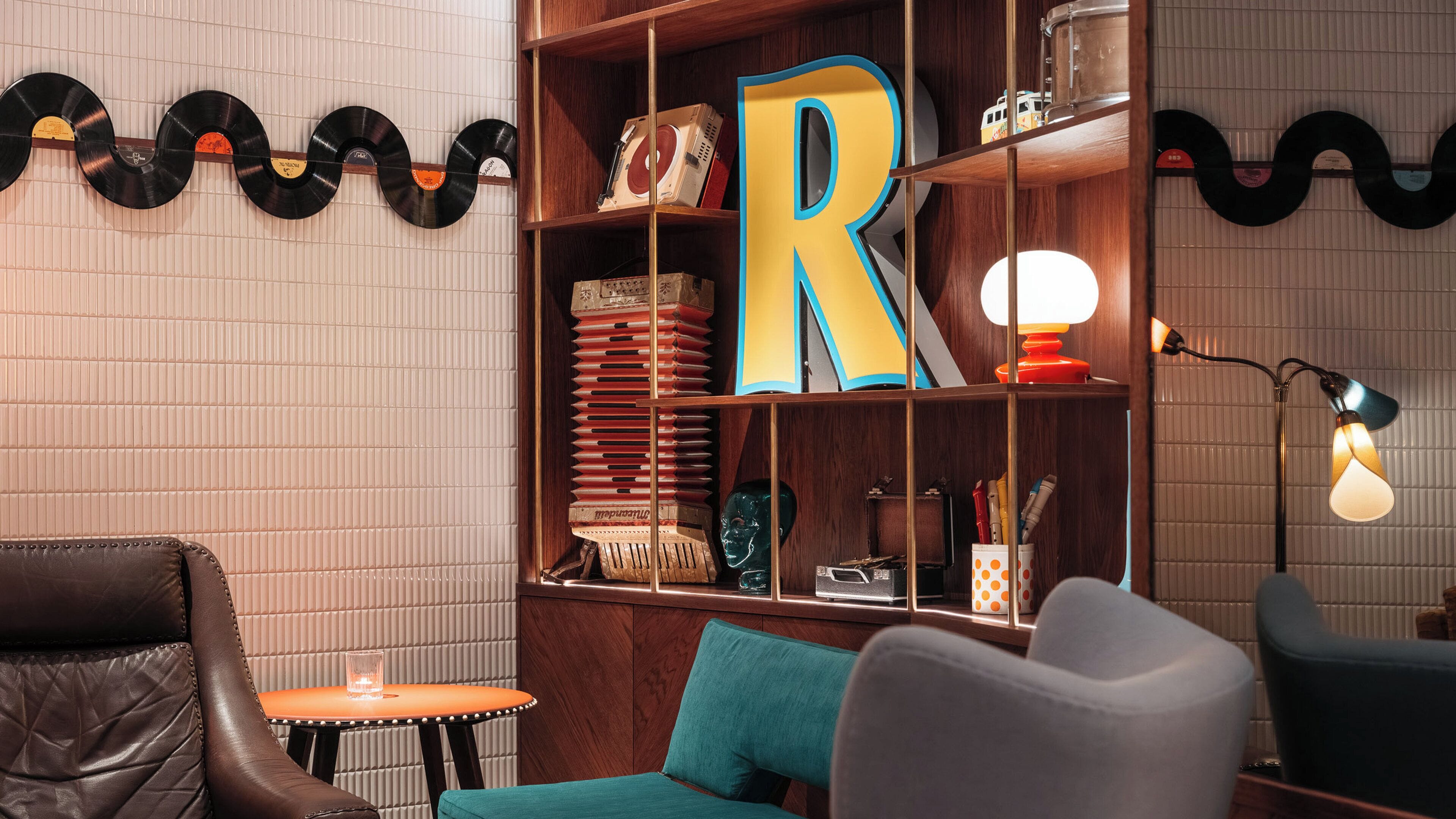 A room with a wooden bookshelf, a large 'R' decoration, vinyl records, and a lamp.