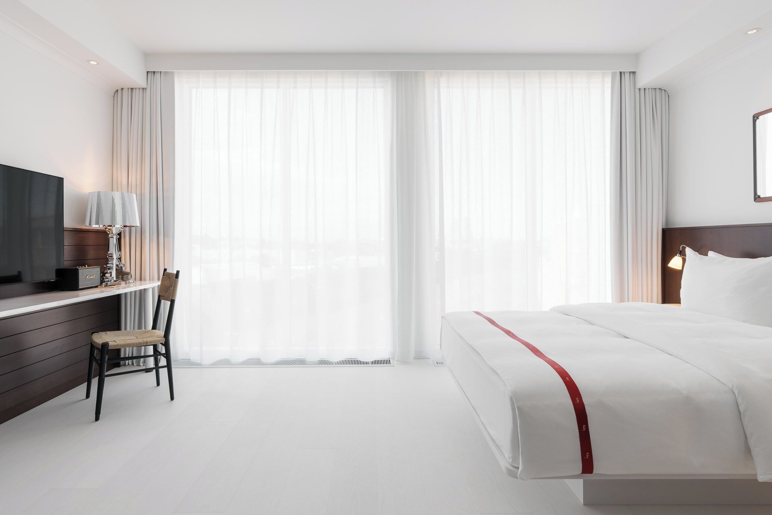 Una habitación de hotel con cama, escritorio, silla, televisor y ventanas amplias con cortinas.