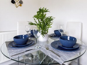 A round glass table with blue-rimmed bowls and wine glasses, a white vase with a plant, and a white chair. - One Bedroom Luxury Apartment (London)