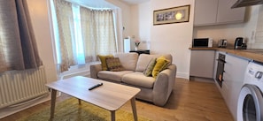 A compact living space with a sofa, a small table, a washing machine, and a kitchenette. - Apartment Sleeps 4 with WiFi (Clacton-on-Sea)