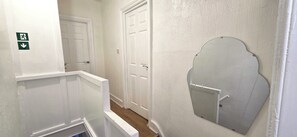 A narrow hallway with white walls and doors, a small mirror on the right wall, and a green emergency exit sign on the left. - Apartment Sleeps 4 with WiFi (Clacton-on-Sea)