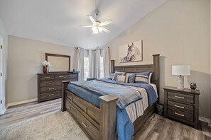 Ein Schlafzimmer mit einem großen Bett, einem Nachttisch mit Lampe, einer Kommode, einem Deckenventilator und einem Fenster mit Vorhängen.