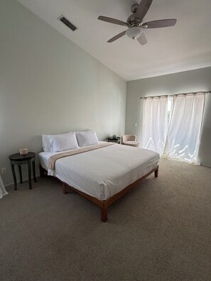 Ein Schlafzimmer mit einem Bett, Deckenventilator, Nachttisch, Stuhl und einem Fenster mit Vorhängen.