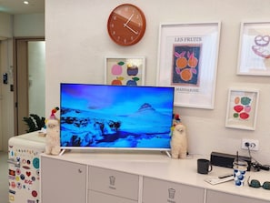 A room with a large flat-screen TV on a white cabinet, a clock on the wall, and framed artwork depicting fruits.