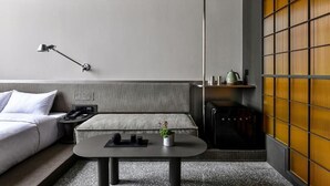 A modern hotel room with a grey sofa, a small round table with a plant, a bedside lamp, and a wall-mounted shelf with a kettle. - NOA HOTEL XIAMEN (Xiamen)