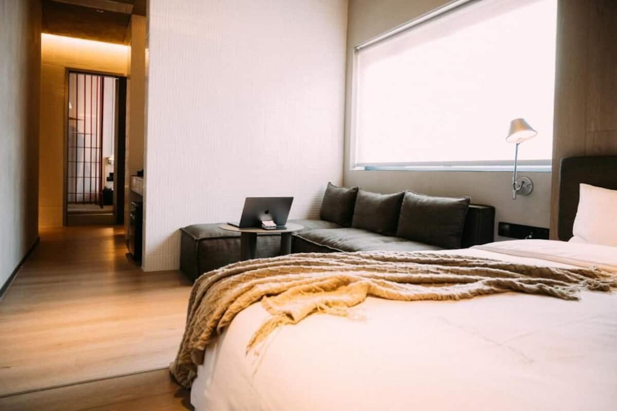 Un dormitorio con cama, un ordenador portátil sobre una mesita de noche, una ventana con vistas y una puerta que da a otra habitación.