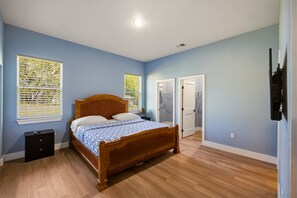 A bedroom with a wooden bed, two windows with blinds, a bedside table, a wardrobe, and a TV mounted on the wall.