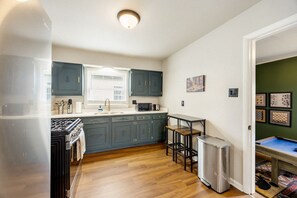 Cocina compacta con estufa a gas, lavamanos y microondas. Incluye un refrigerador pequeño y una mesa con dos sillas. Los armarios son azul oscuro y hay una ventana sobre el lavamanos.