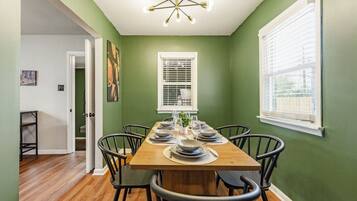 A dining area with a wooden table set for six, surrounded by black chairs, a ceiling light fixture, and a window with blinds.