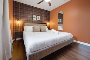 A neatly made bed with white linens and a grey headboard in a room with wooden flooring and plaid wallpaper.