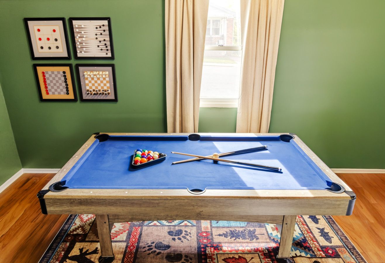 Ein Billardtisch mit Kugeln und Spielstangen, ein gemustertes Teppich und gerahmte Schachsets an der Wand.