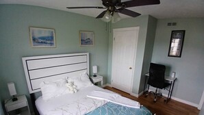 A neatly made bed with a white headboard, a ceiling fan, two framed pictures on the wall, a white door, and a small table with a chair.