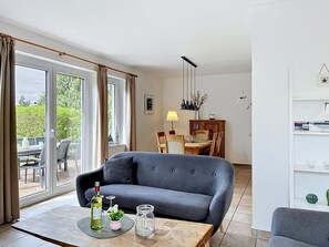 Ein modernes Wohnzimmer mit grauem Sofa, Esstisch und Stühlen, großen Fenstern mit Vorhängen und Blick ins Grüne.