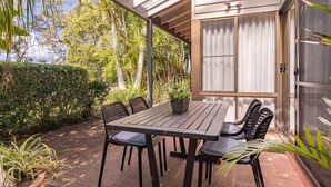 Un patio al aire libre con una mesa y sillas, rodeado de follaje y un edificio con ventanas amplias.