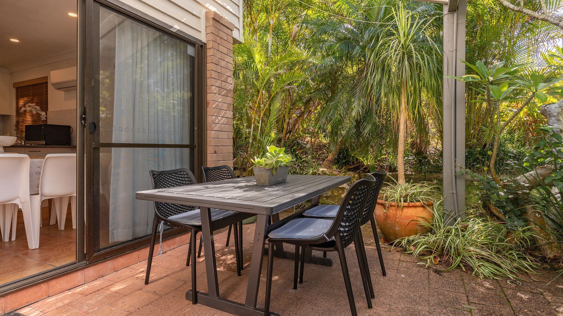 An outdoor patio with a table and chairs, surrounded by lush greenery and a brick wall.