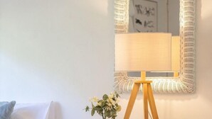 Una habitación con una mesa consola de mimbre blanco, un jarrón pequeño con flores, un taburete de madera, un sofá blanco y un espejo montado en la pared.