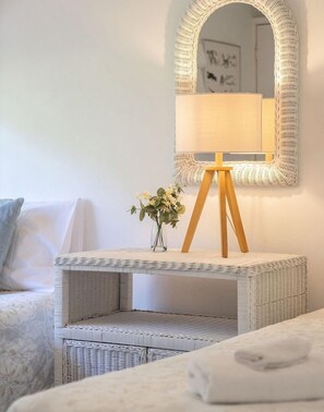 Ein Zimmer mit einer weißen Korbtischkonsole, einer kleinen Vase mit Blumen, einem Holzschemel, einer weißen Couch und einem Wandspiegel.