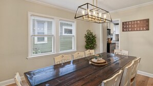A dining area with a wooden table, white chairs, a hanging light fixture, and a wall sign listing cities. - Big Group Getaway Near OSU with 6BR, 8 Beds, 5-Car Parking (Columbus)