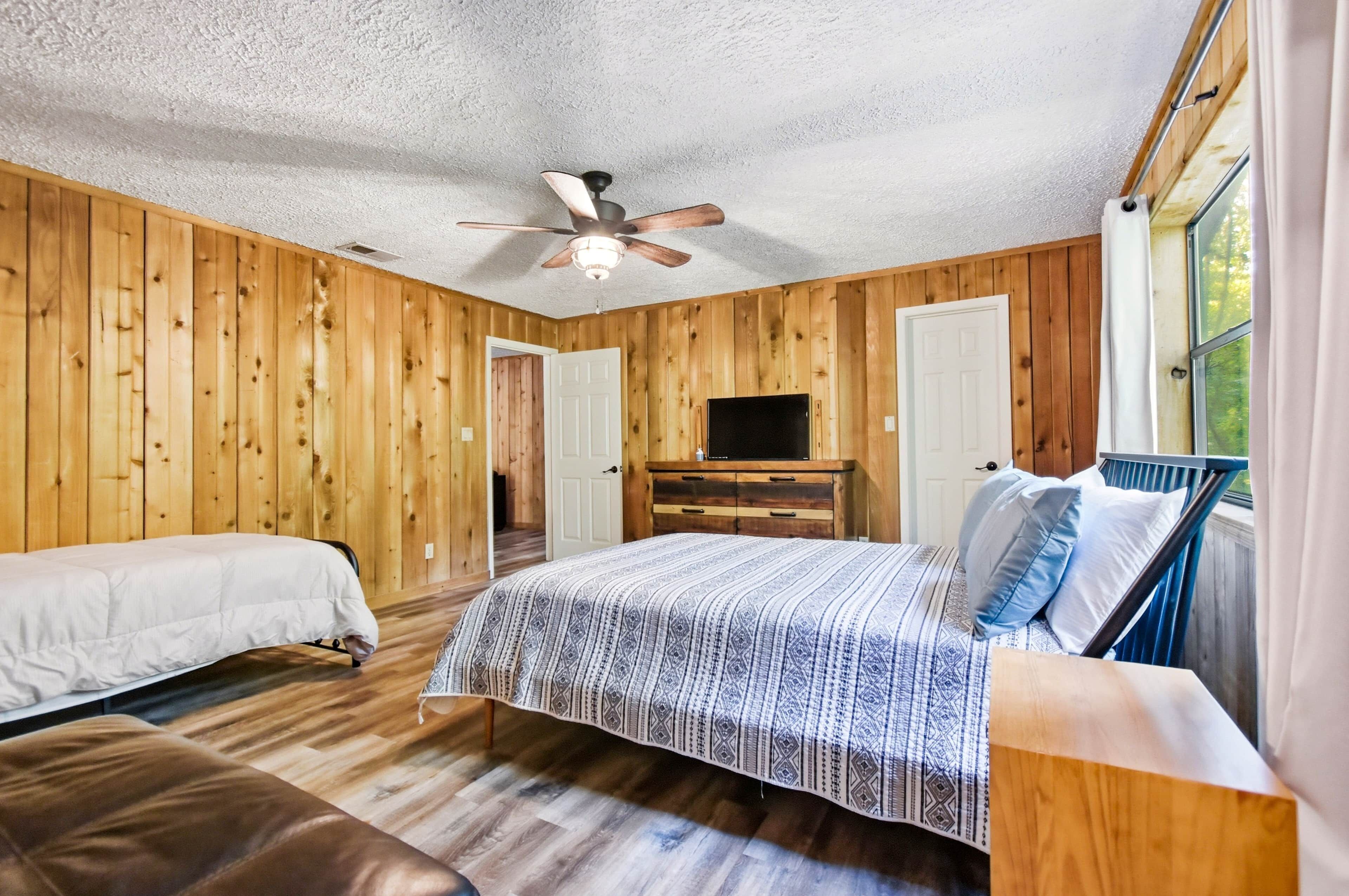 Une pièce aux murs en bois, avec un ventilateur de plafond, un lit recouvert d’une courtepointe à motifs, un banc, une commode et un téléviseur.