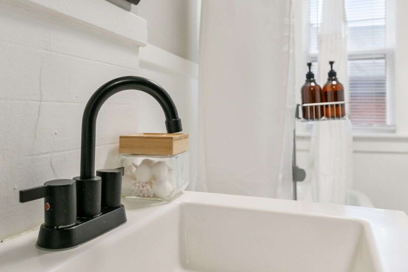 A bathroom with a white sink, a black faucet, a glass jar with white flowers, and a window with blinds.