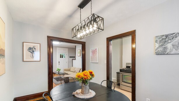 A dining area with a round table, chairs, a vase with flowers, a hanging light fixture, and a kitchen visible through an open door.
