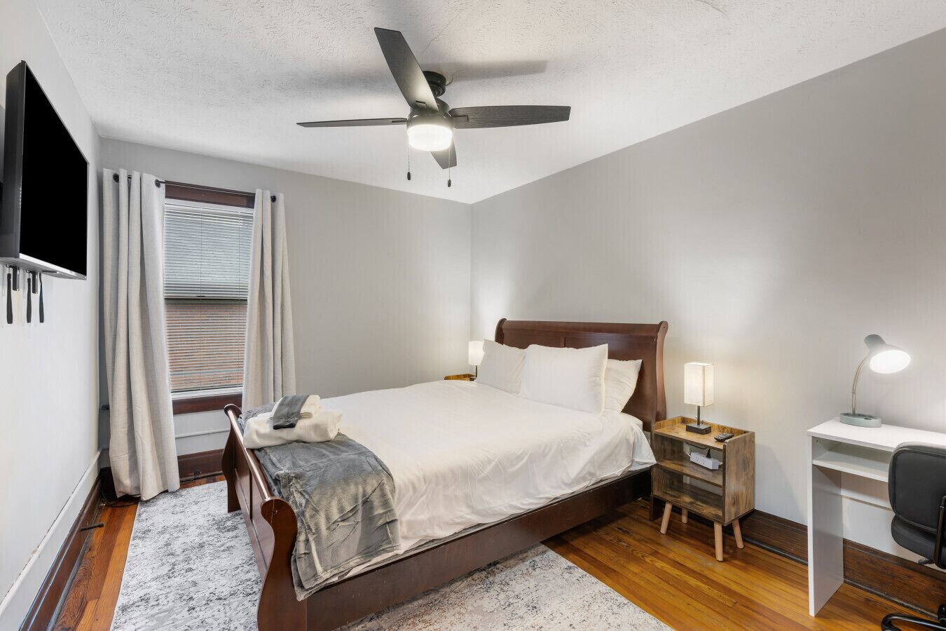 Un dormitorio con cama, mesitas de noche, escritorio, silla, ventilador de techo y televisor.