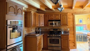 Interior de una cabaña de madera con cocina equipada, electrodomésticos integrados, ventilador de techo y un dormitorio visible con cama y cómoda.