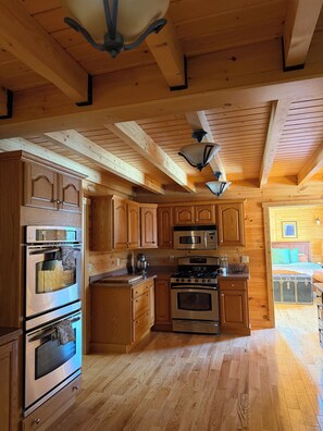 A wooden cabin interior with a kitchen featuring built-in appliances, a ceiling fan, and a visible bedroom with a bed and dresser.