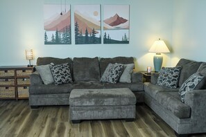 Ein graues Ecksofa mit gemusterten Kissen, ein passendes Ottomanen, ein Beistelltisch mit Lampe sowie Wandbilder mit Bergen und Bäumen.