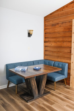 Ein Eckzimmer mit einer Holzwand, einer blau gepolsterten Bank, einem Holztisch mit einer Schachtel und einer Fernbedienung sowie einer Wandleuchte.