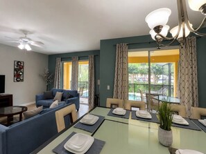 A modern living room with a sofa, dining table set for four, and a balcony with a pool view. - Paradise Palm Home Stay (Kissimmee)