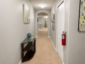 A hallway with a glass-top console table, a fire extinguisher mounted on the wall, and artwork on the walls. - Paradise Palm Home Stay / Near parks (Kissimmee)