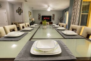 A dining room with a long table set for a meal, a television displaying 'NETFLIX' in the background, and a ceiling fan above. - Paradise Palm Home Stay / Near parks (Kissimmee)